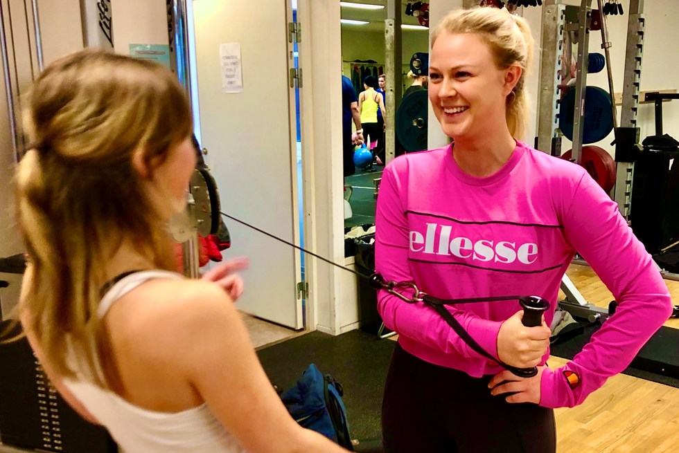 Leende kvinna tränar på ett gym med en person framför sig.